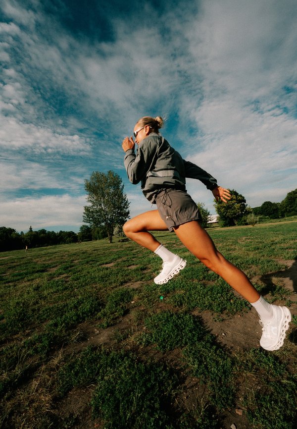 Kobieta w sportowym stroju i okularach przeciwsłonecznych biegnąca w powietrzu po zielonym polu pod częściowo pochmurnym niebieskim niebem.