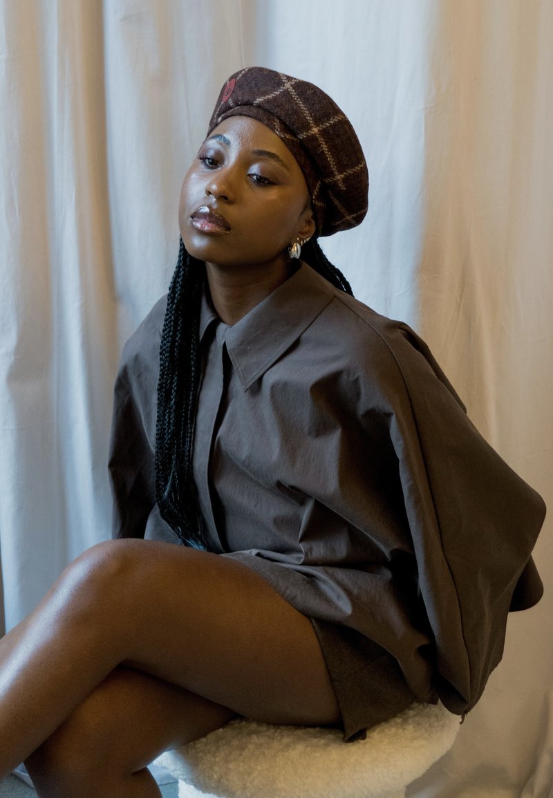 Brwon oversized button-up shirt and matching shorts, paired with a patterned brown beret. The outfit features a smooth texture and relaxed fit, set against a light backdrop.