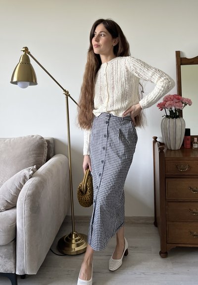 Suéter blanco de punto con patrón texturizado, falda midi de cuadros en blanco y negro con botones, zapatos blancos de tejido y un bolso marrón.