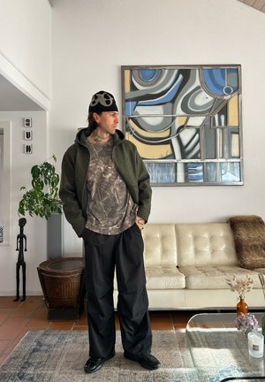 Hombre con gorro con estampado, chaqueta verde oscuro y pantalones negros, de pie en una sala de estar con arte abstracto en la pared y un sofá de cuero beige.