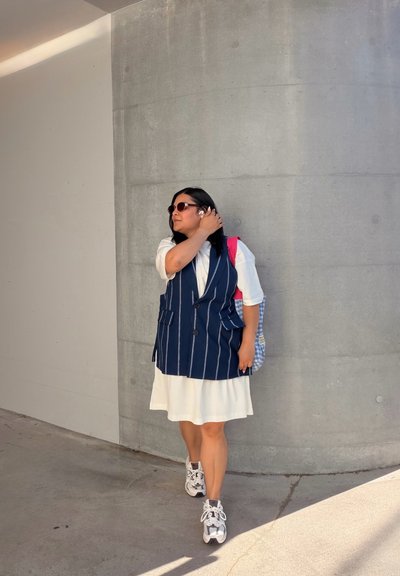 Mujer con gafas de sol, vestido blanco, chaleco azul marino a rayas y zapatillas deportivas, ajustándose las gafas de sol mientras está de pie junto a una pared curva de hormigón.