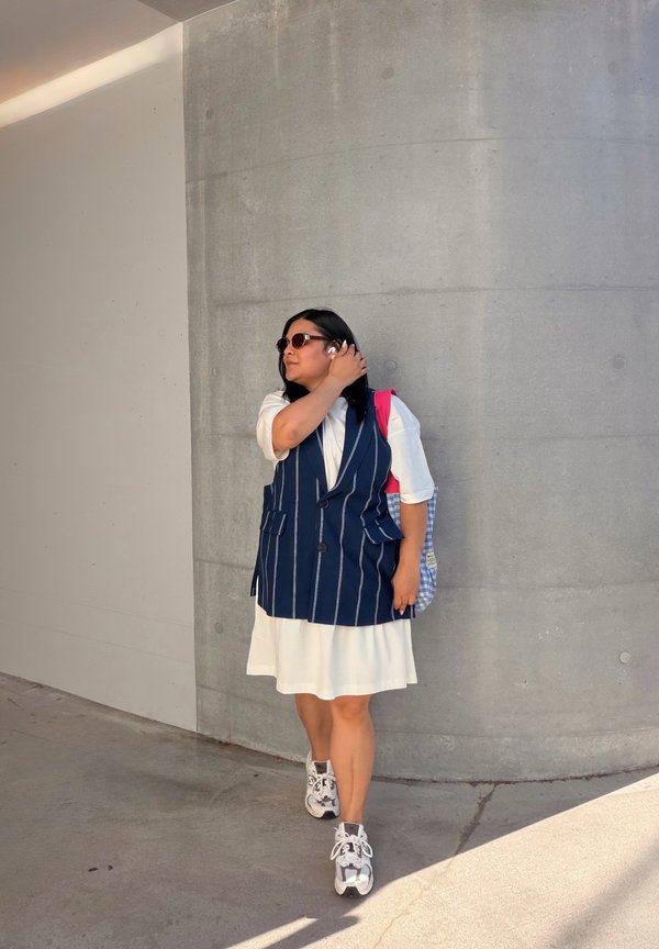 Femme portant des lunettes de soleil, une robe blanche, un gilet bleu marine à fines rayures et des baskets, ajustant ses lunettes de soleil tout en se tenant contre un mur en béton courbé.