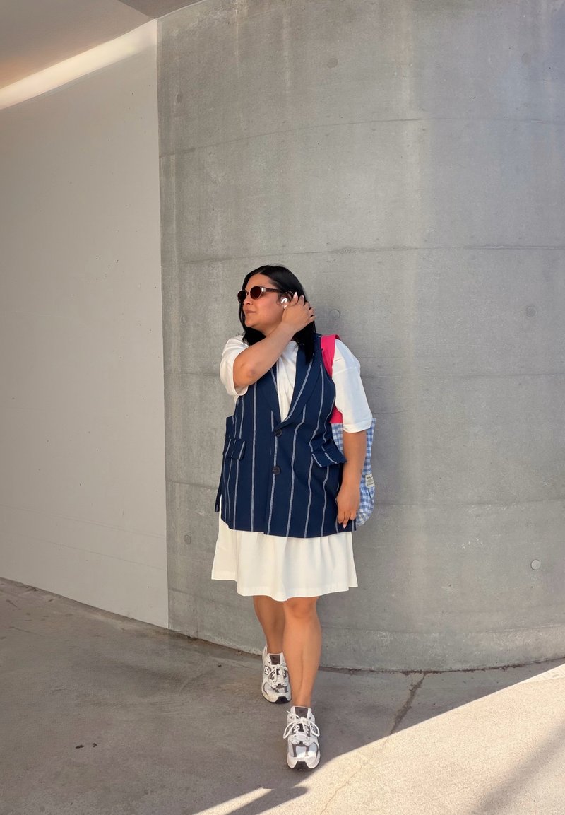 Frau mit Sonnenbrille, weißem Kleid, marineblau gestreifter Weste und Turnschuhen, die ihre Sonnenbrille anpasst, während sie gegen eine gebogene Betonwand lehnt.