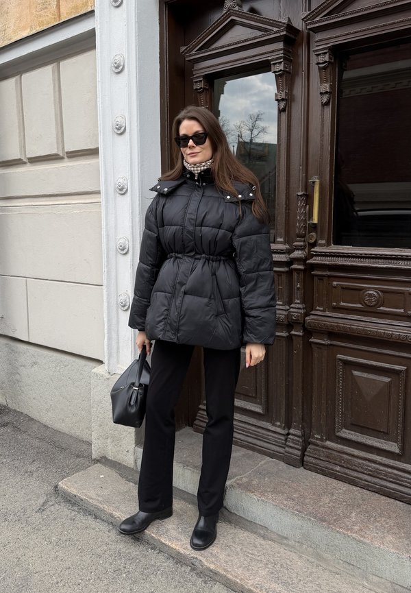 Femeie îmbrăcată într-o jachetă neagră matlasată, pantaloni negri, cizme, ochelari de soare și cu o geantă neagră, stând lângă o ușă mare de lemn maro, ornamentată.