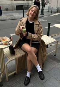 Trench-coat beige, pull noir et short. Assis avec une boisson verte. Mocassins noirs et chaussettes blanches. Petite table avec bols à proximité.