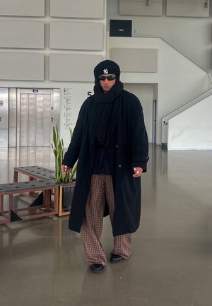 Un abrigo largo negro sobre un conjunto negro, pantalones marrones con patrón, una sudadera con capucha negra y gafas de sol. De pie en el interior cerca de ascensores y vegetación.