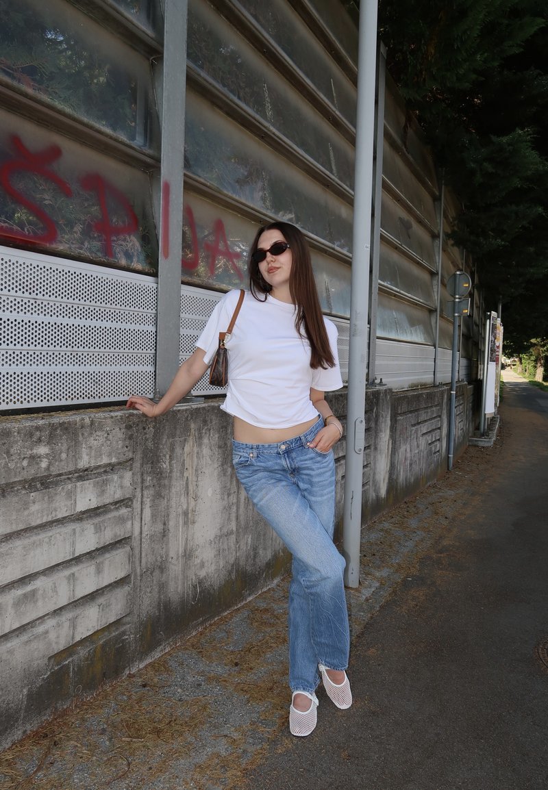 Jeune femme portant des lunettes de soleil, un t-shirt blanc et un jean, appuyée contre un mur en béton tagué sur un trottoir en ville.