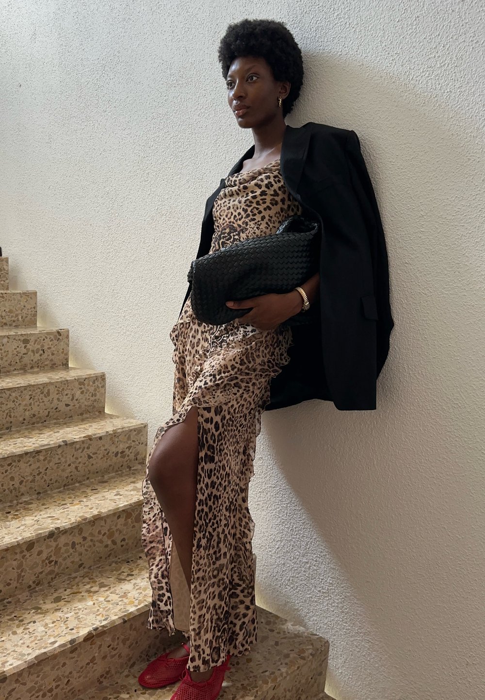 Vestido de estampado de leopardo com uma fenda alta, blazer oversized preto e uma clutch preta texturizada. Sapatos vermelhos elaboradamente tecidos completam o visual.