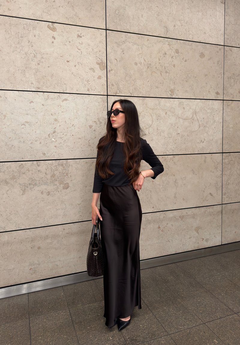 Donna con lunghi capelli scuri che indossa top nero, gonna di raso nera, scarpe nere appuntite, occhiali da sole e porta una borsa nera davanti a un muro piastrellato.