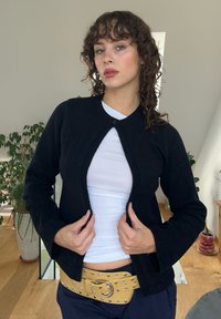 Μαύρο cardigan με στρογγυλή λαιμόκοψη, μπροστινό κλείσιμο με κουμπιά, ribbed άκρες και μακριά μανίκια, φορεμένο πάνω από ένα λευκό τοπ και κίτρινη ζώνη.