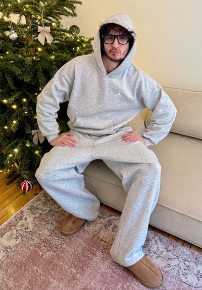 Joven con pantalones deportivos grises y sudadera con capucha, que lleva gafas y zapatillas, sentado en un sofá cerca de un árbol de Navidad decorado con luces.
