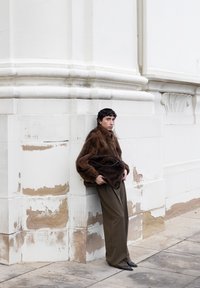 Person, der læner sig mod en hvid, vejrslidt stenvæg iført en brun pelsjakke, olivengrønne bukser og holder en matchende brun pels taske.