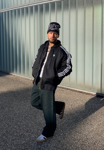 Hombre con chaqueta negra de Adidas, gorro y zapatillas posa con las manos en los bolsillos frente a una pared de vidrio corrugado al aire libre.