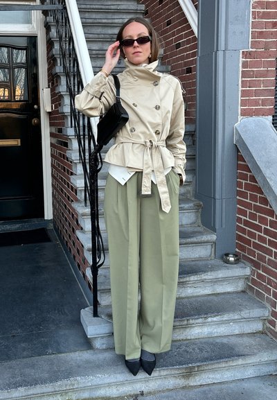 Gabardina beige con botones de doble botonadura, ceñida en la cintura. Pantalones verdes de pierna ancha, tacones negros de punta y un bolso negro pequeño.