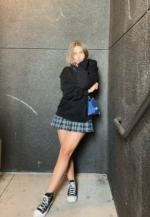 Sudadera negra de gran tamaño, falda corta de cuadros con acentos azules y zapatillas de caña alta en blanco y negro. Fondo de pared texturizada gris.