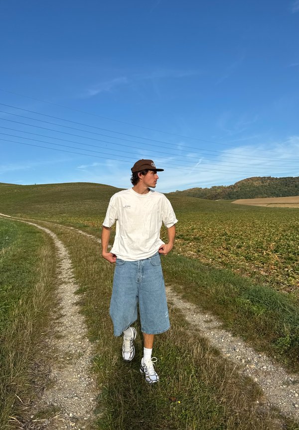 Joven con camisa blanca, pantalones cortos de mezclilla y gorra marrón caminando por un camino de tierra a través de campos verdes bajo un cielo azul claro.