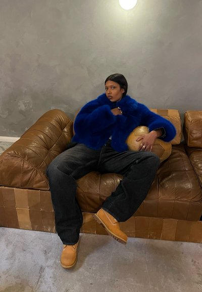 Chaqueta de pelo sintético azul, pantalones de mezclilla oscuros y botas de tobillo beige, sentado en un sofá de cuero marrón, sosteniendo un objeto esférico.