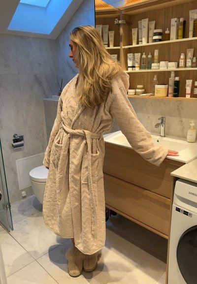 Bata de baño acolchada beige con capucha, zapatillas de lana, de pie junto a un lavabo en un baño moderno con estanterías de madera llenas de productos de skincare.