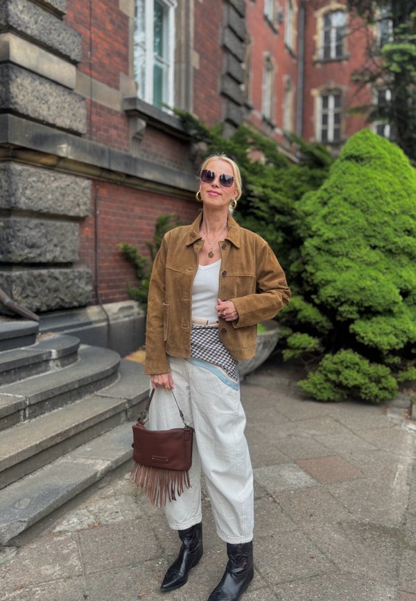 Blonde vrouw met een bruine jas, witte broek, zwarte laarzen, zonnebril en een bruine handtas met franjes staat bij stenen treden en groene struiken.