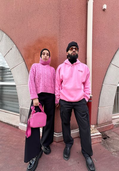 Dos personas se encuentran de pie contra una pared texturizada de color rosa. La mujer lleva un suéter grueso de punto de cable rosa y pantalones anchos negros, sosteniendo un bolso rosa. El hombre lleva una sudadera rosa, pantalones negros y un gorro.