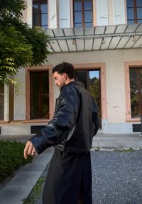 Uomo che indossa una giacca di pelle nera e pantaloni neri mentre cammina all'esterno di un edificio con ampie finestre e vegetazione nelle vicinanze.