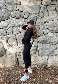 Sudadera deportiva negra y leggings con acentos dorados. Zapatillas blancas. De pie contra una pared de piedra texturizada, con hojas esparcidas abajo.