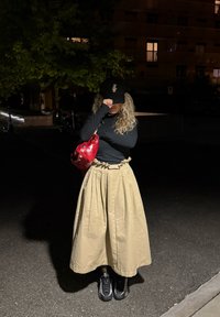 Lang beige skjørt med volangliv, svart langermet topp, svart baseballcaps og rød håndveske; svarte joggesko fullfører looken.