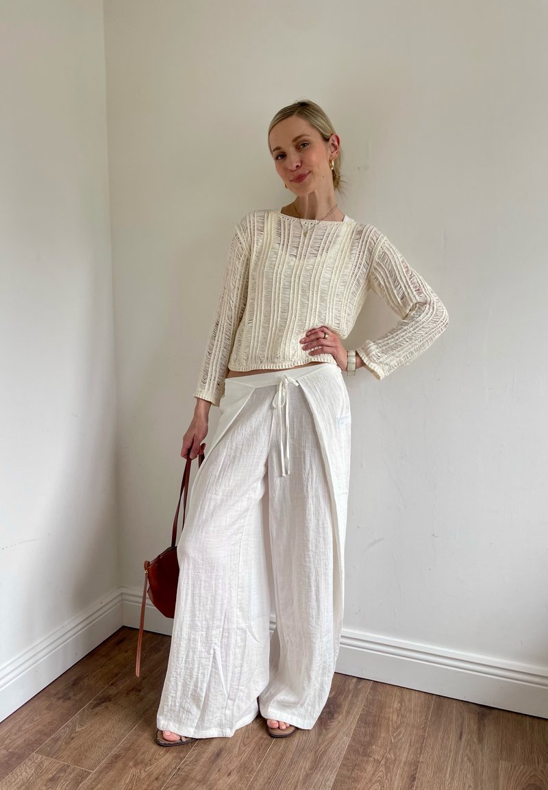 Femme portant un haut blanc texturé à manches longues et un pantalon blanc à jambes larges, debout avec une main sur la hanche et tenant un sac brun dans l'autre.