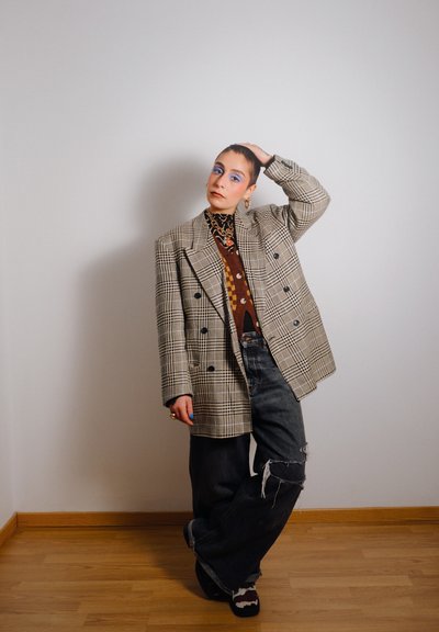 Blazer de cuadros de doble botonadura en beige y negro, superpuesto sobre una camisa marrón con estampado. Combinado con unos vaqueros oscuros desgastados y un calzado singular.