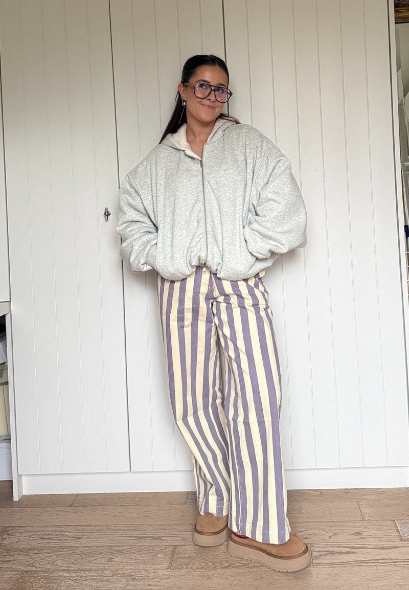 Sweatshirt cinza claro oversized com fecho de correr, calças de perna larga às riscas em creme e lavanda, e sapatos castanhos sem atilho com sola grossa.