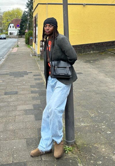 Una persona se apoya contra una farola, vestida con un blazer gris, un suéter naranja, unos vaqueros holgados azul claro y botas de tobillo marrones, sosteniendo una bolsa negra.