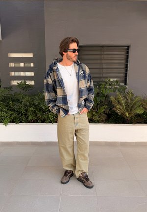 Hombre con gafas de sol, chaqueta a cuadros sobre una camisa blanca, pantalones beige y zapatillas marrones, de pie sobre un suelo de baldosas junto a una pared con plantas.