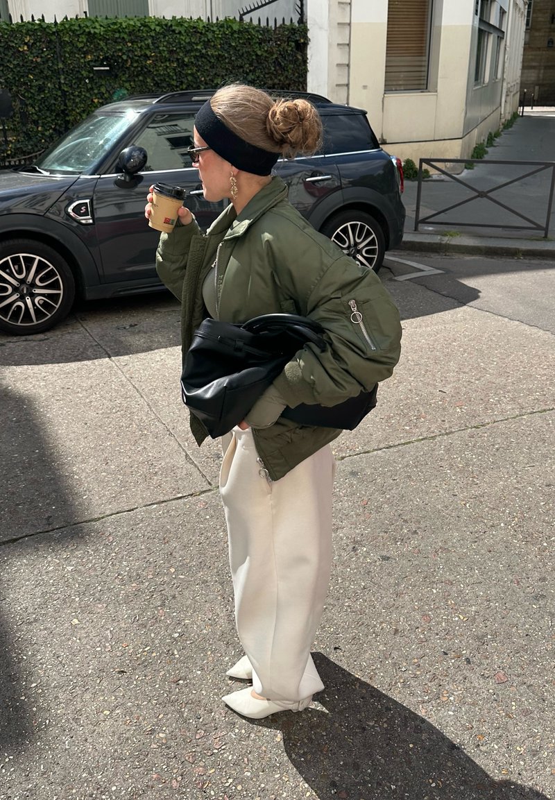Femme en veste verte et pantalon blanc, tenant un sac noir et buvant dans un gobelet de café à emporter dans une rue ensoleillée.