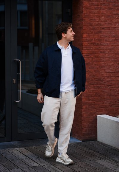 Chaqueta bomber navy sobre una camiseta polo blanca, combinada con pantalones de lino claros y zapatillas beige. El entorno presenta ladrillo rojizo y suelo de madera.