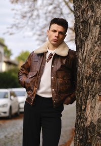 Giacca di pelle marrone con colletto in pelliccia, due tasche frontali e design vintage. Indossata sopra un maglione crema e pantaloni neri. Ambientazione naturale.