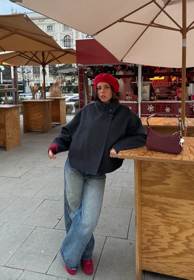 Chaqueta de lana gris con cuello alto y detalles de botones, combinada con jeans de pierna ancha y zapatos rojos. Un boina y bolso rojos completan el conjunto.