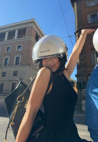 Γυναίκα με μαύρο αμάνικο φόρεμα φορώντας λευκό κράνος Michael Kors χαμογελά και σηκώνει ένα χέρι σε εξωτερικό χώρο σε ηλιόλουστο αστικό περιβάλλον.