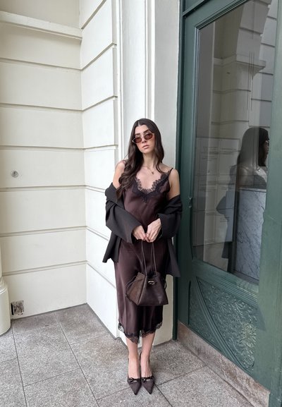 Mujer con cabello largo y oscuro, usando gafas de sol, vestido negro con encaje en el borde, blazer oversize, tacones puntiagudos y sosteniendo un bolso pequeño.