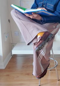 Pessoa sentada num banco a ler um livro, vestindo calças de treino rosa da Adidas, meias amarelas, sapatos de couro castanho e uma jaqueta de ganga azul.