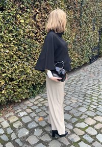 Top nero stile poncho, pantaloni beige a gamba larga e scarpe nere con tacco blocco, con una piccola borsa nera con manico.