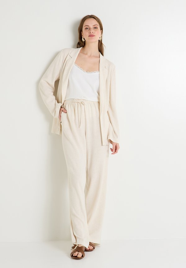 Blazer de lino beige con corte relajado, combinado con pantalones de pierna ancha a juego y una camiseta sin mangas con borde de encaje blanco. Sandalias marrones completan el look.