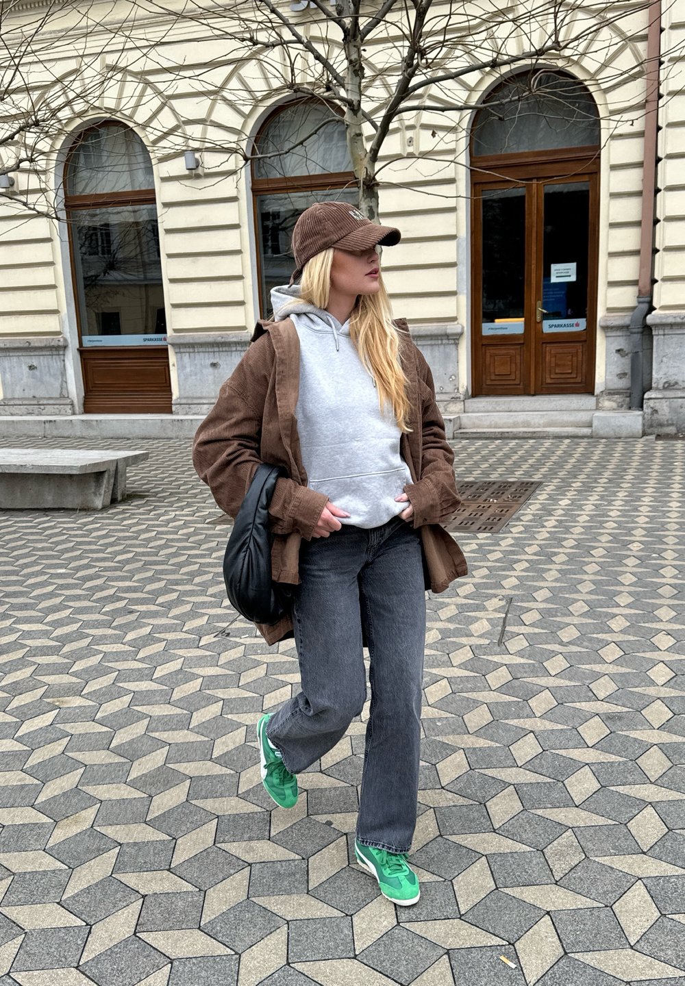 Abrigo marrón con capucha sobre un sudadera gris, jeans grises y zapatillas verdes. Bolsa de hombro negra. Pavimento de adoquines y edificio al fondo.