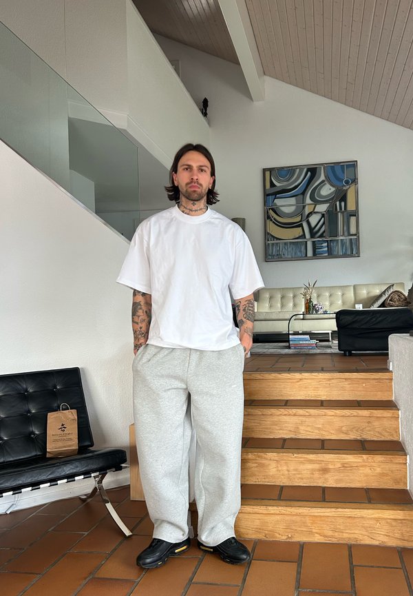 Mann med tatoveringer iført hvit oversized t-skjorte, grå joggebukser og svarte joggesko som står i en moderne stue ved siden av trekonstruksjon av trapp.