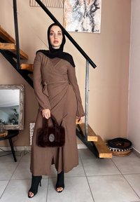 Καφέ ριγέ φόρεμα τύπου περιτύλιξης με μακριά μανίκια και διάκοσμο με κορδόνια, συνδυασμένο με μια σκούρα γούνινη τσάντα και μαύρες ανοιχτές μπότες, τοποθετημένο μπροστά από μια σκάλα.