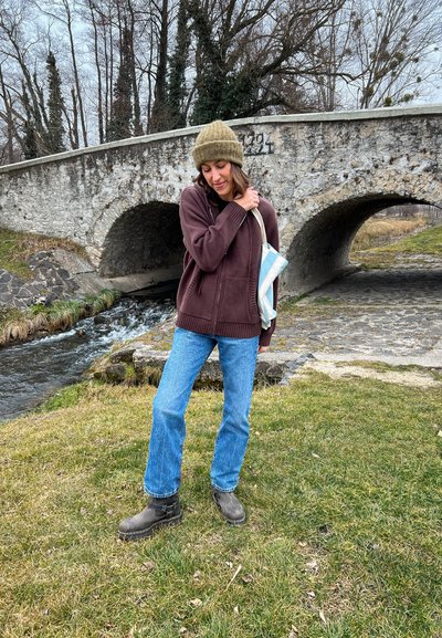 Sudadera con cremallera marrón, vaqueros azul claro, botas gris de tobillo. El modelo sostiene un bolso a rayas. El fondo presenta un puente de piedra y un arroyo.