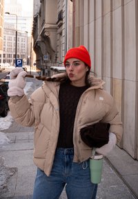 Jeune femme portant un bonnet rouge, une doudoune beige et des gants pelucheux, tenant une tresse et un gobelet vert sur un trottoir enneigé en ville.