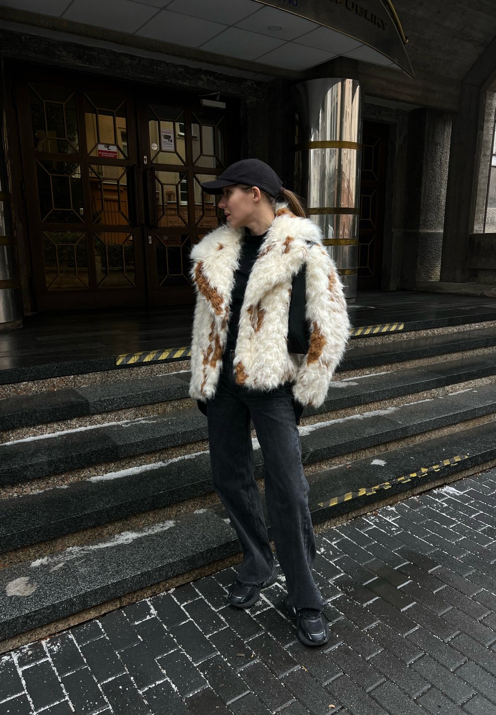 Femme portant une casquette noire, un manteau en fourrure blanche et brune, un pantalon noir et des chaussures, debout sur des marches en pierre à l'extérieur d'un bâtiment avec des portes vitrées.
