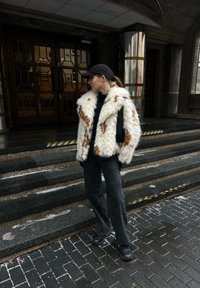 Femme portant une casquette noire, un manteau en fourrure blanche et brune, un pantalon noir et des chaussures, debout sur des marches en pierre à l'extérieur d'un bâtiment avec des portes vitrées.