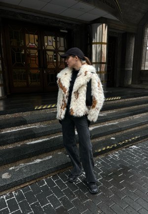Mujer con gorra negra, abrigo de piel blanca y marrón, pantalones y zapatos negros, de pie sobre unos escalones de piedra frente a un edificio con puertas de vidrio.