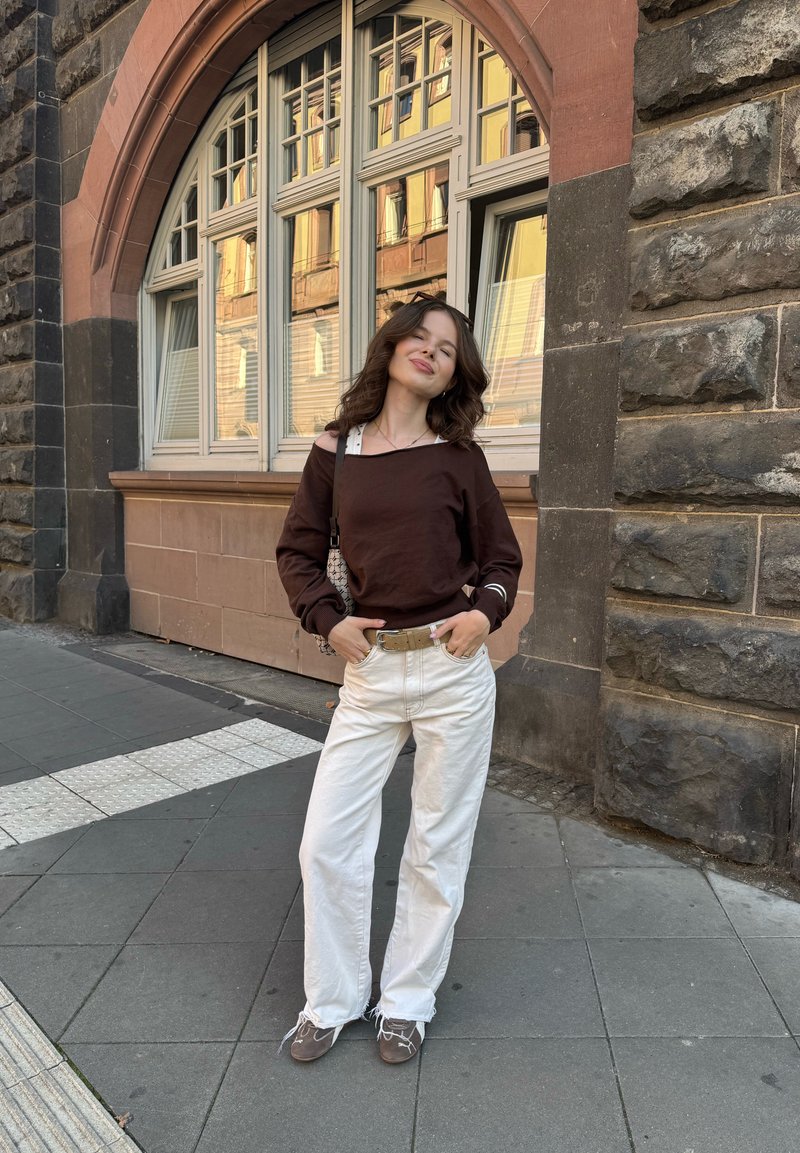 Haut en long-sleeve marron, style off-shoulder, pantalon large crème, et chaussures grises avec lacets, se tenant contre un mur en pierre.